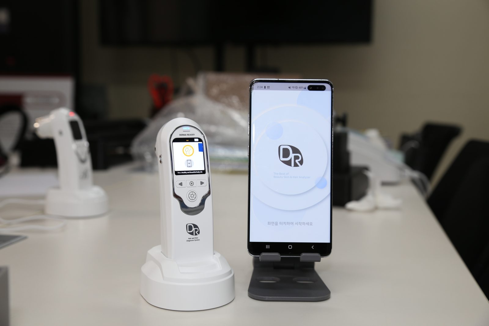충전 거치대에 놓인 DERMA READER 디바이스와 앱 시작 화면을 띄운 스마트폰